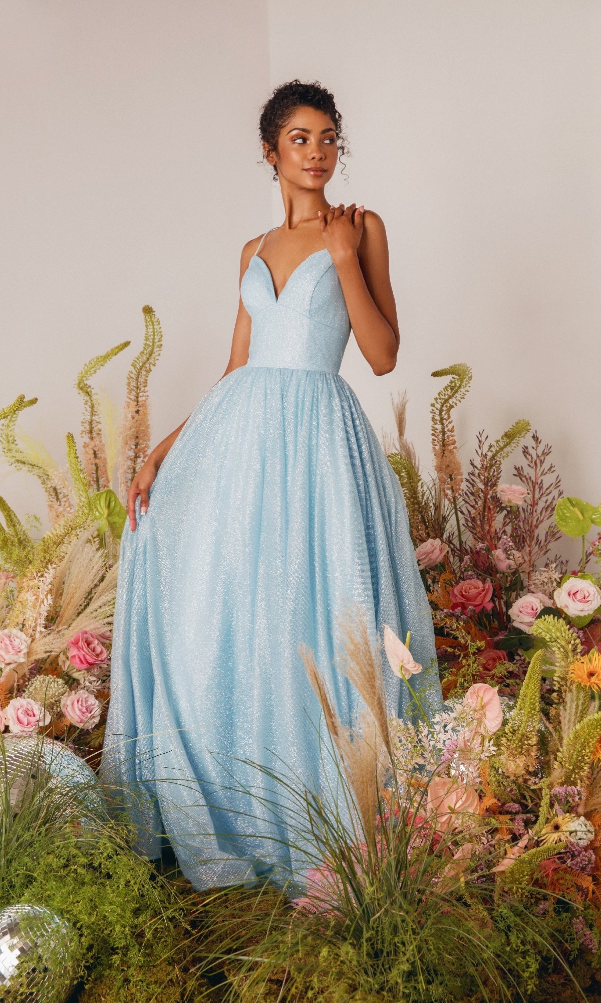 A woman in the Strappy-Back Glitter Prom Ball Gown: Eureka 9009, a light blue sparkly evening dress with thin straps, stands among flowers and grasses in a softly lit room.